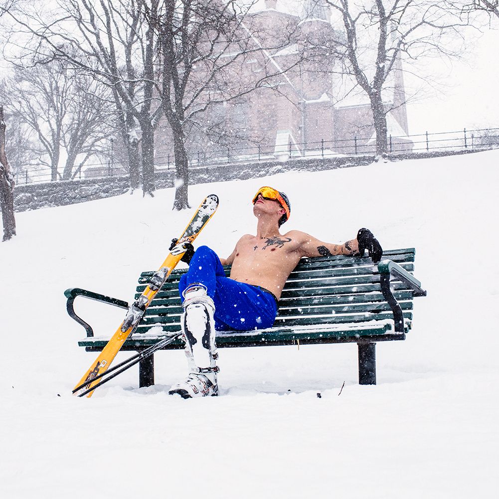 A man sitting shirtless on a bench in a snowy park, wearing åäö’s The Pants in Brilliant Blue, with skis resting by his side.