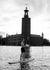 A man standing relaxed on a paddleboard, wearing åäö’s Deep Blue Shorts, with Stockholm City Hall in the background.