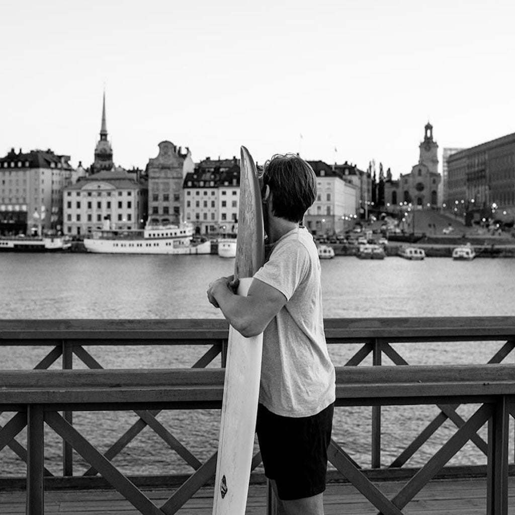 A man in åäö Shorts standing in front of water and boats with arms around surfboard.