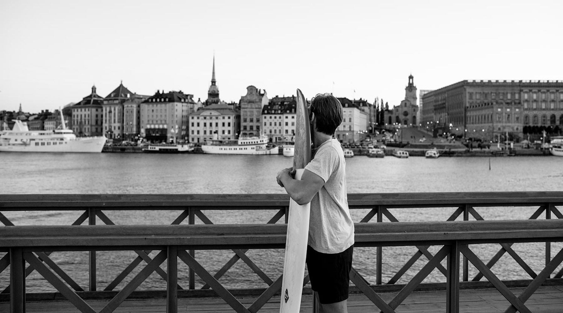 A man in åäö Shorts standing in front of water and boats with arms around surfboard.