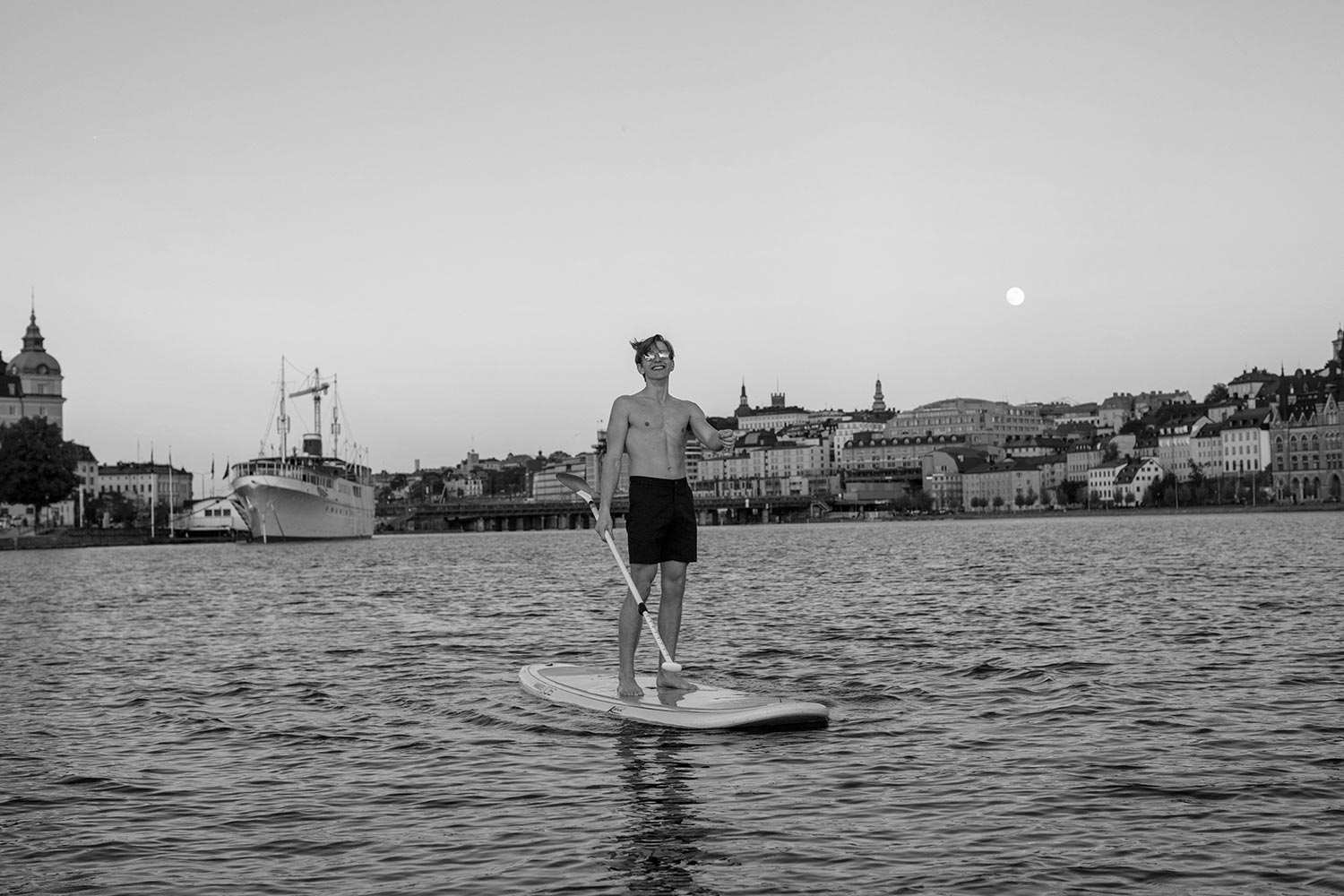 A man standing relaxed on a paddleboard, wearing åäö’s Deep Blue Shorts, with Stockholm’s skyline in the background