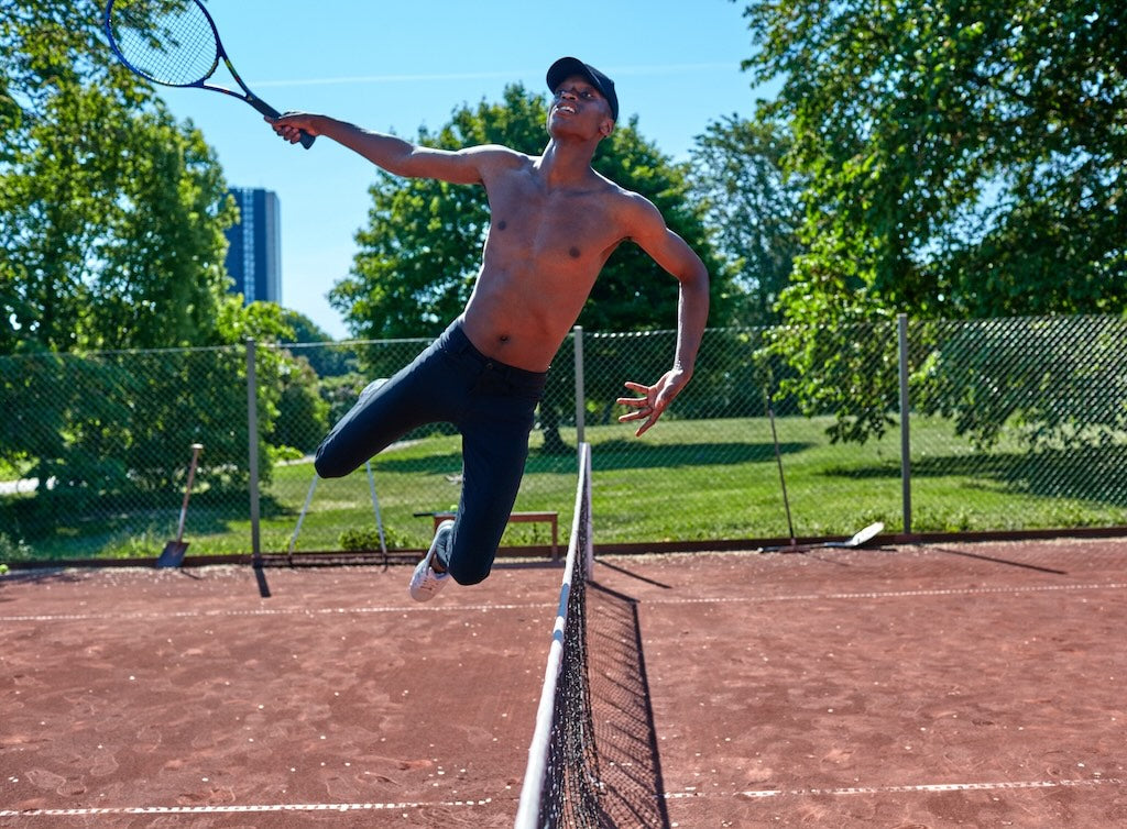 A man jumping over the net on a tennis court, wearing The Pants by åäö, showcasing their flexibility and design.