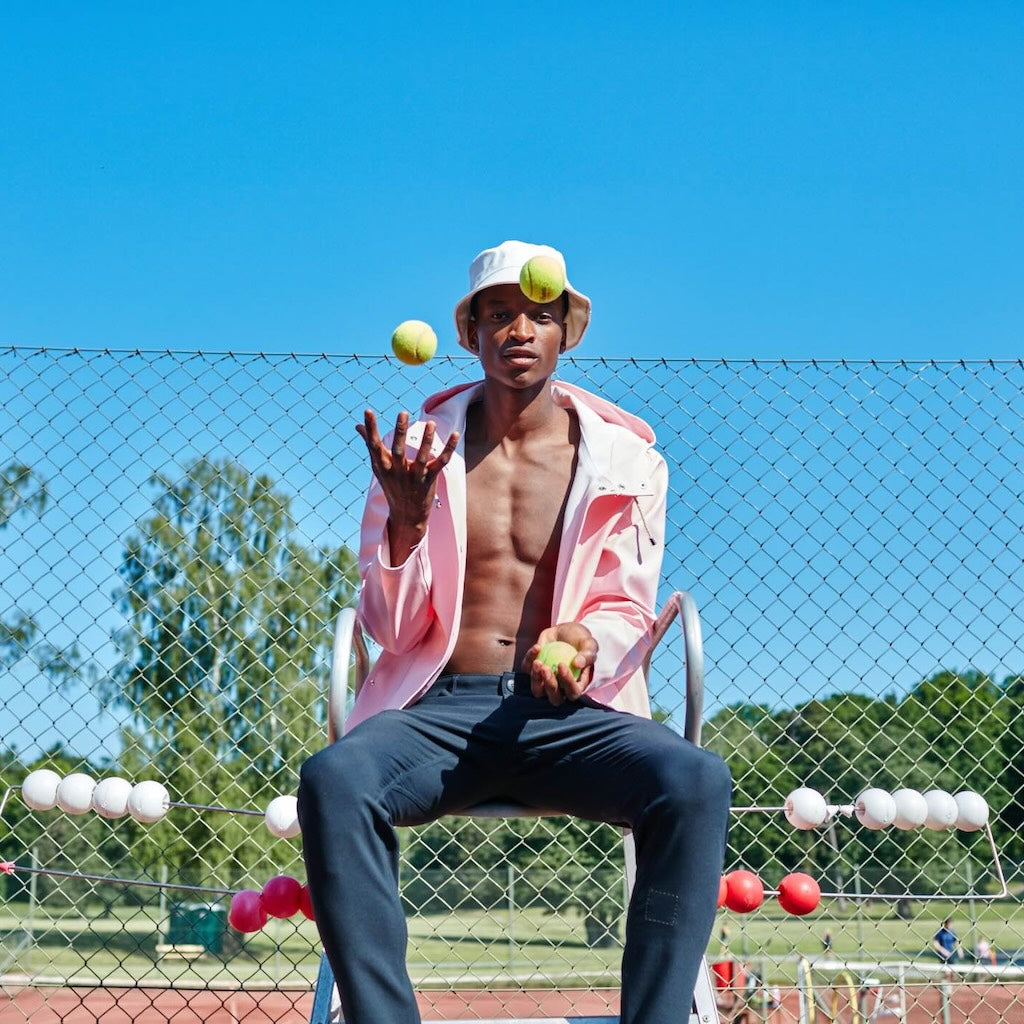 A man in The Pants by åäö in Blue Black sits confidently on an umpire chair on a tennis court, juggling three tennis balls.