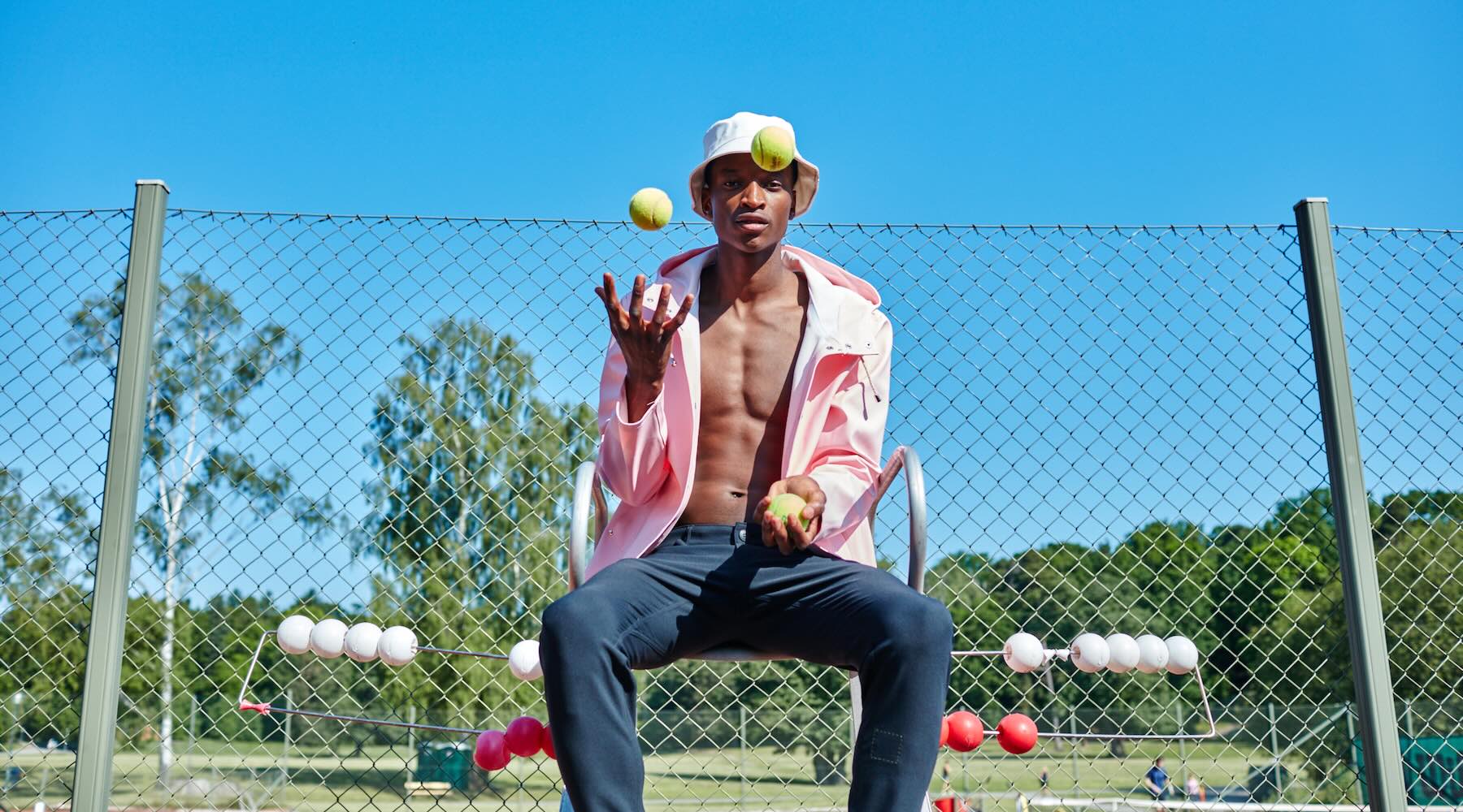 A man in The Pants by åäö in Blue Black sits confidently on an umpire chair on a tennis court, juggling three tennis balls.