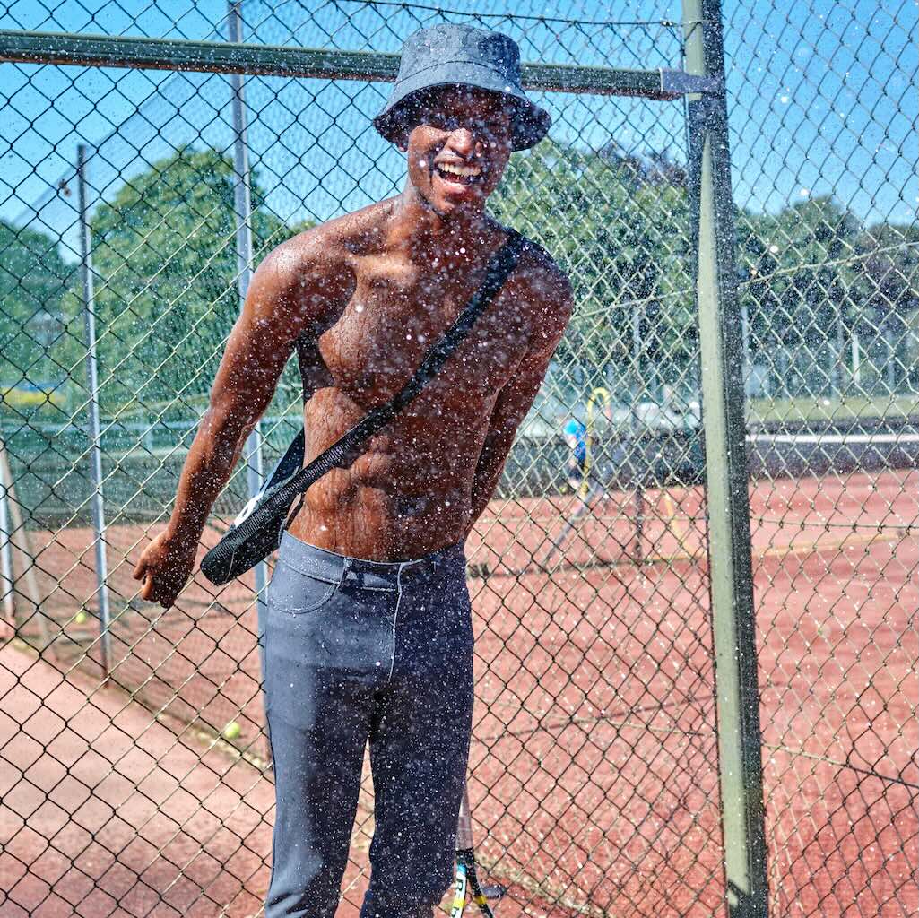 A man standing by the fence on a tennis court, wearing The Pants by åäö, laughing and enjoying the moment.