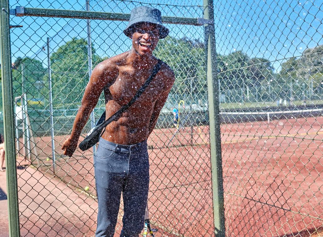 A man standing by the fence on a tennis court, wearing The Pants by åäö, laughing and enjoying the moment.