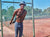 A man standing by the fence on a tennis court, wearing The Pants by åäö, laughing and enjoying the moment.