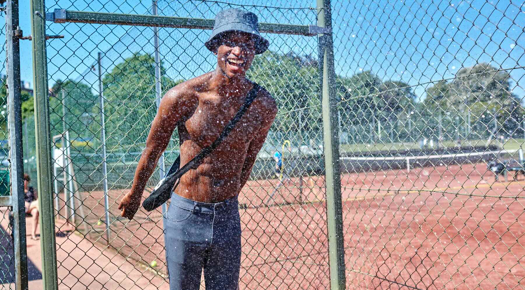 A man standing by the fence on a tennis court, wearing The Pants by åäö, laughing and enjoying the moment.