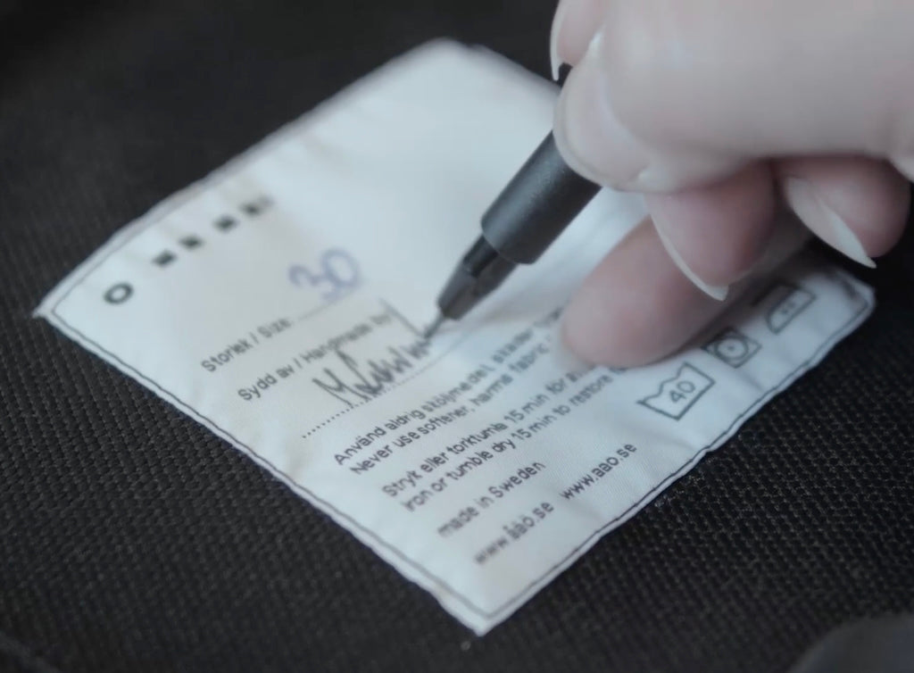 Close-up of a seamstress’s hand signing the size badge inside åäö’s Pants, showcasing attention to craftsmanship and detail.